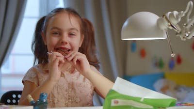 Happy Little Girl is Eating a Chocolate Bar Sitting at the Table.