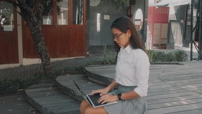 Asian Attractive Young Woman Wearing Glasses Working at Her Laptop on the Street.