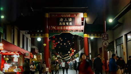 A busy street at night adorned with chinese lights has several people walking under a traditional Chinese door.