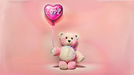 A cute teddy bear holding a heart-shaped balloon with the word love on it walks over a pink backdrop.
