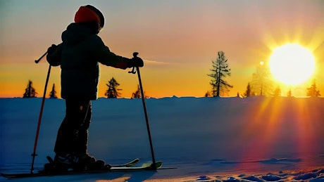 A kid in skiing gear is walking on the snow at dusk.