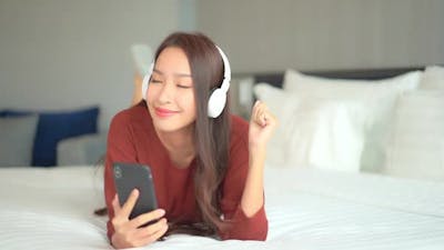 A close-up of a young woman prone on a big comfy bed wearing headphones and enjoying the music from .