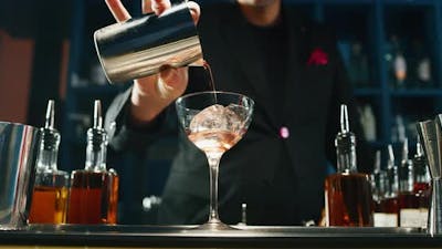 A professional bartender is preparing an alcoholic cocktail with ice cubes to customers at the bar.